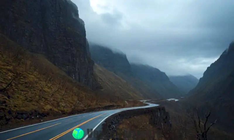 Carretera peligrosa en un paisaje montañoso, con acantilados y neblina, rodeada de árboles muertos y señales de advertencia